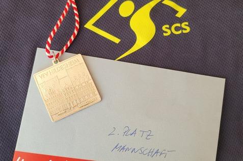 Medaille mit rot-wei&szlig;er Schnur, Umschlag mit Aufschrift &bdquo;2. Platz Mannschaft&ldquo; auf einem dunklen Trikot.