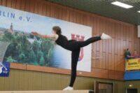 Turnerin in schwarzem Outfit balanciert auf einem Schwebebalken in einer Turnhalle. Zuschauer im Hintergrund.