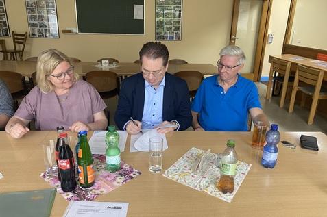 Drei Personen sitzen an einem Tisch mit Getränken und Unterlagen; eine Person unterschreibt ein Dokument.