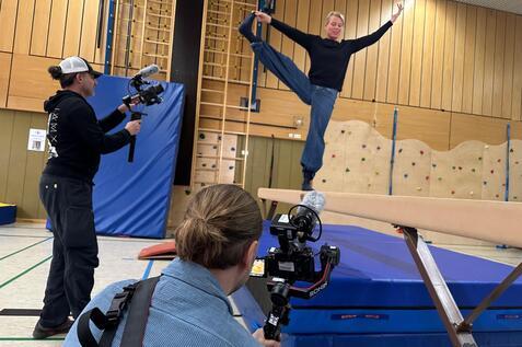 Eine Person balanciert auf einem Schwebebalken, w&auml;hrend zwei Kameraleute filmen. Hintergrund mit Kletterwand und Matten.