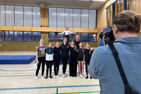 Gruppe von sechs Sportlerinnen in Sportkleidung posiert in einer Turnhalle, w&auml;hrend ein Fotograf ein Foto aufnimmt.