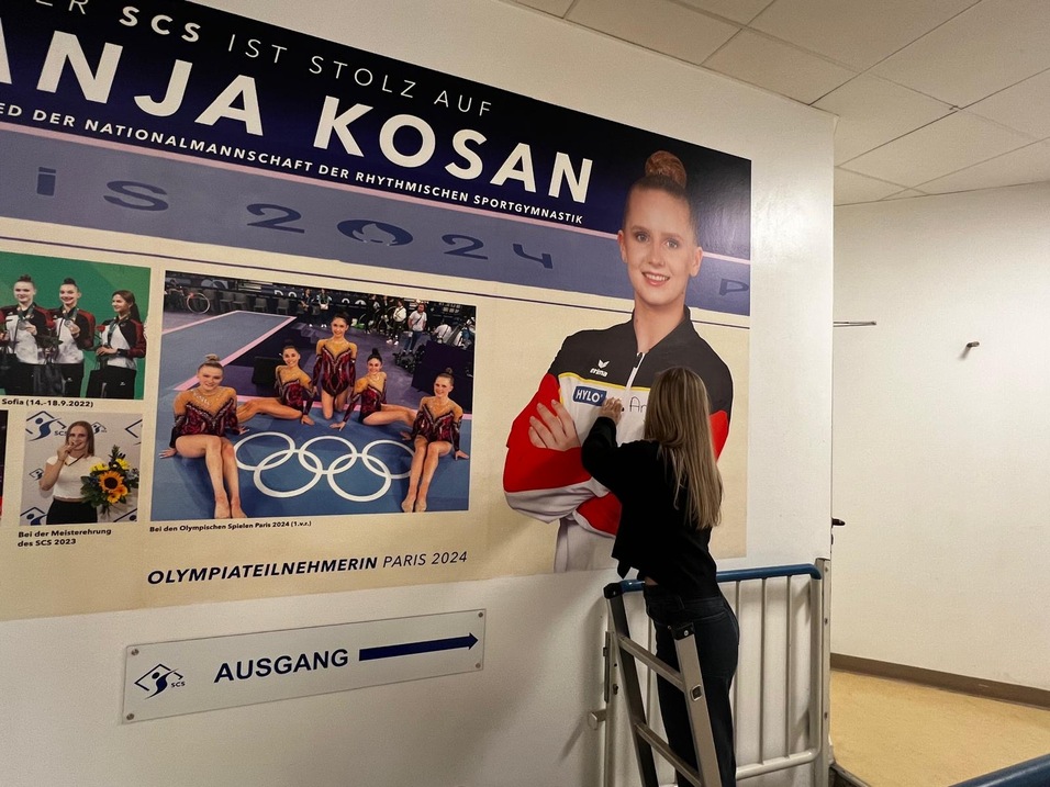 Frau auf einer Leiter, die ein Bild von Anja Kosan, einer Sportlerin, an einer Wand anbringt. Olympische Ringe sichtbar.
