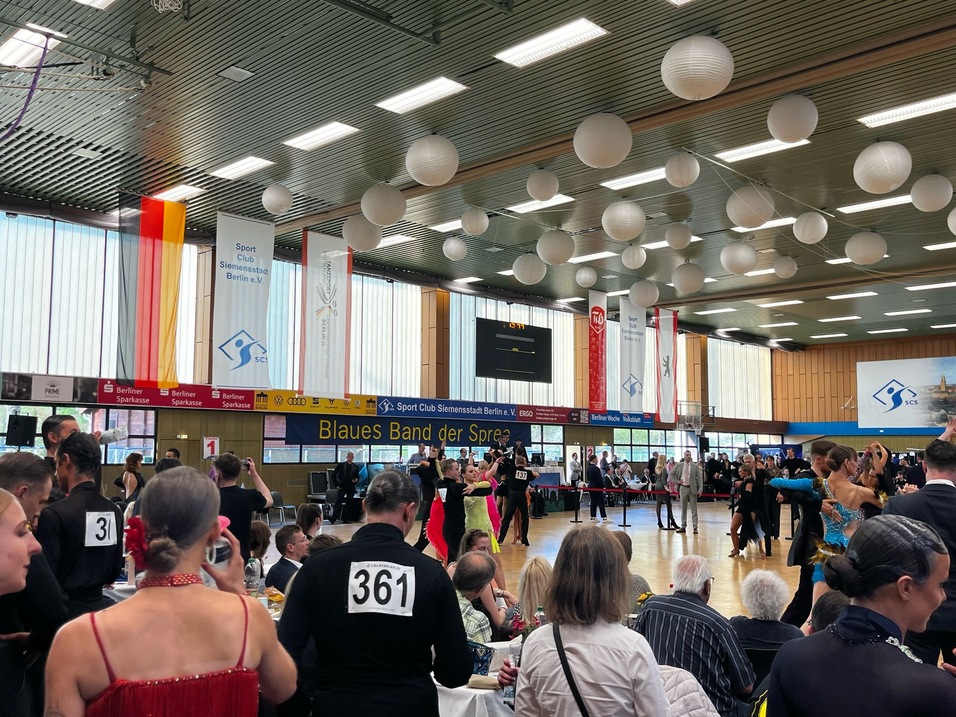 Tanzturnier in einer Halle mit Teilnehmern und Zuschauern, dekoriert mit weißen Lampions und bunten Bannern.