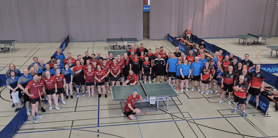 Gruppenfoto von Tischtennisspielern in Sportkleidung auf einer Sporthalle mit Tischtennisplatten im Hintergrund.