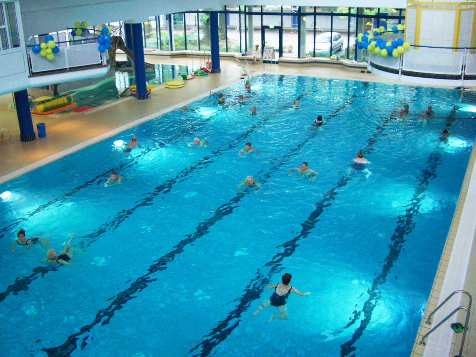 Innenansicht eines Schwimmbads mit blauen Wasser; mehrere Personen schwimmen, rutschen und entspannen.