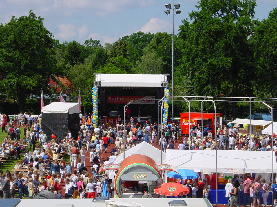 Menschenmenge auf einem Festgelände vor einer Bühne, umgeben von Ständen und Bäumen, bei sonnigem Wetter.