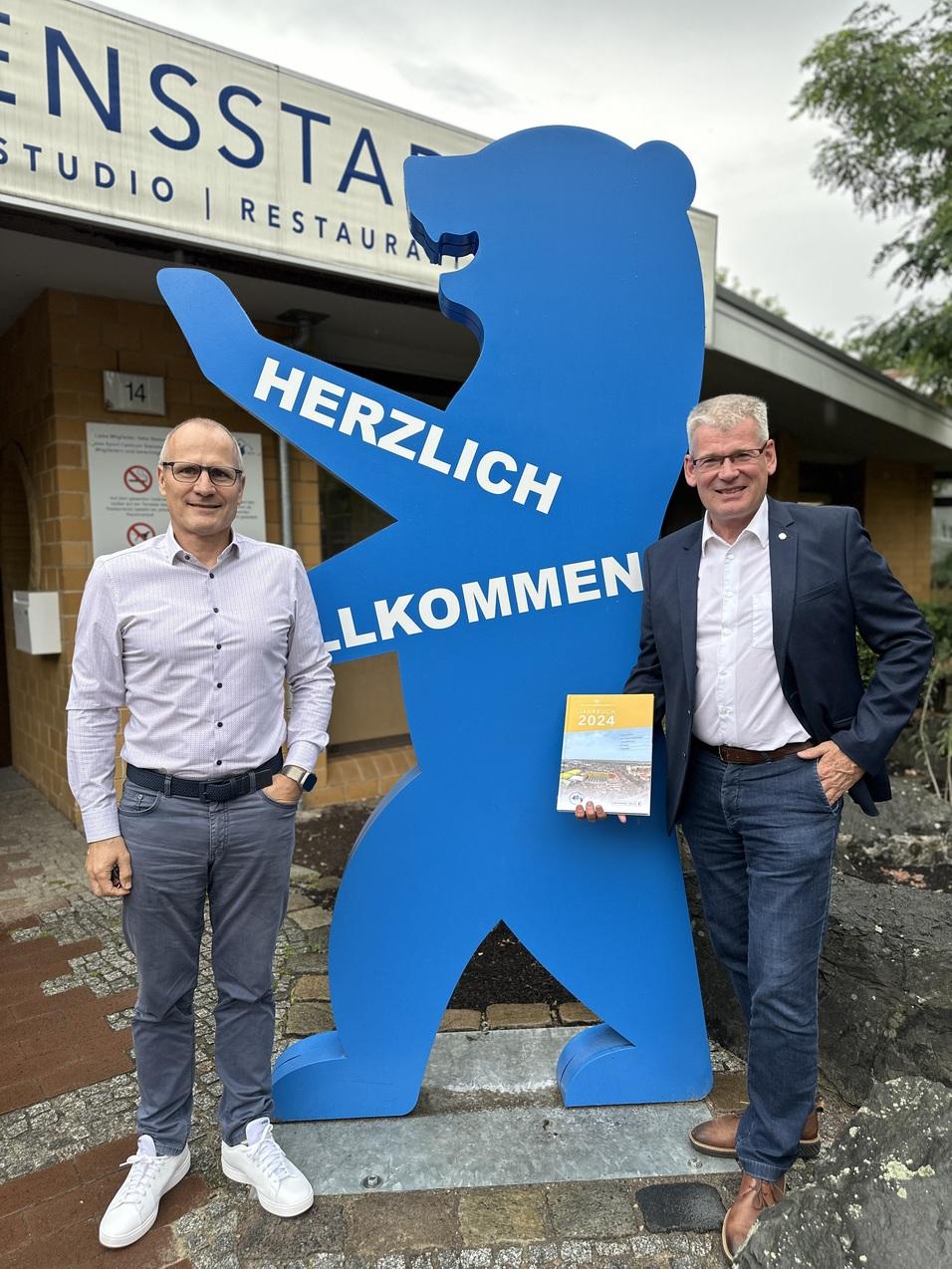 Zwei Männer posieren neben einem blauen Bären mit der Aufschrift „HERZLICH WILLKOMMEN“ vor einem Restaurant.