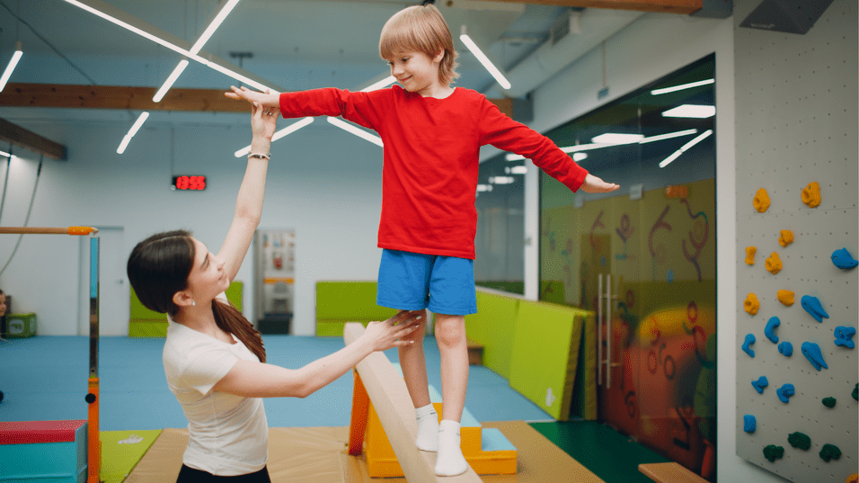 Junge balanciert auf einem Schwebebalken, während eine Trainerin ihn unterstützt, in einer Turnhalle mit Kletterwand.