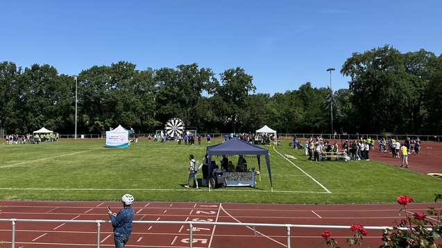 Veranstaltung auf einem Sportplatz mit St&auml;nden, Besuchern und einem gro&szlig;en Gl&uuml;cksrad unter blauem Himmel.