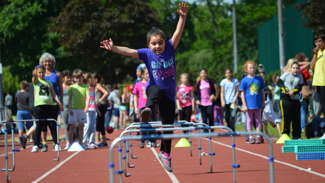 M&auml;dchen in lila T-Shirt springt &uuml;ber H&uuml;rden auf einem Sportplatz, umgeben von anderen Kindern und Zuschauern.