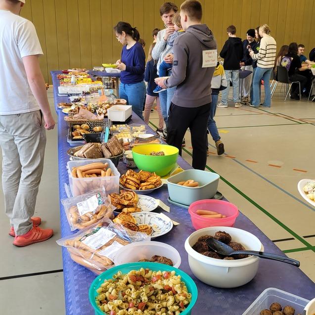 Buffet mit verschiedenen Speisen, unter anderem Salate, Snacks und Wurst, w&auml;hrend G&auml;ste sich unterhalten.