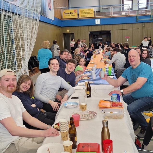 Gruppenszene bei einem Essen in einer Sporthalle, mit mehreren Personen an langen Tischen und Essen auf den Tischen.