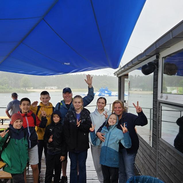 Gruppe von acht Personen steht unter einem blauen Pavillon an einem Gew&auml;sser, einige zeigen Victory-Zeichen.