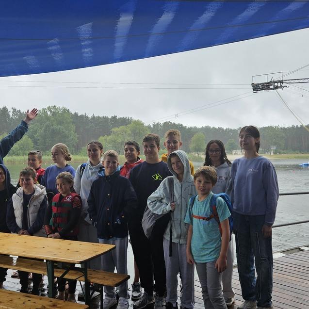 Gruppe von Jugendlichen und Erwachsenen unter einem blauen Sonnenschutz am Wasser, mit Wassersportger&auml;ten im Hintergrund.
