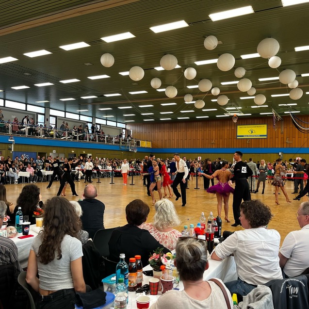 Tanzpaare f&uuml;hren synchron T&auml;nze auf einem Wettbewerb auf, Zuschauer betrachten das Geschehen in einer gro&szlig;en Halle.