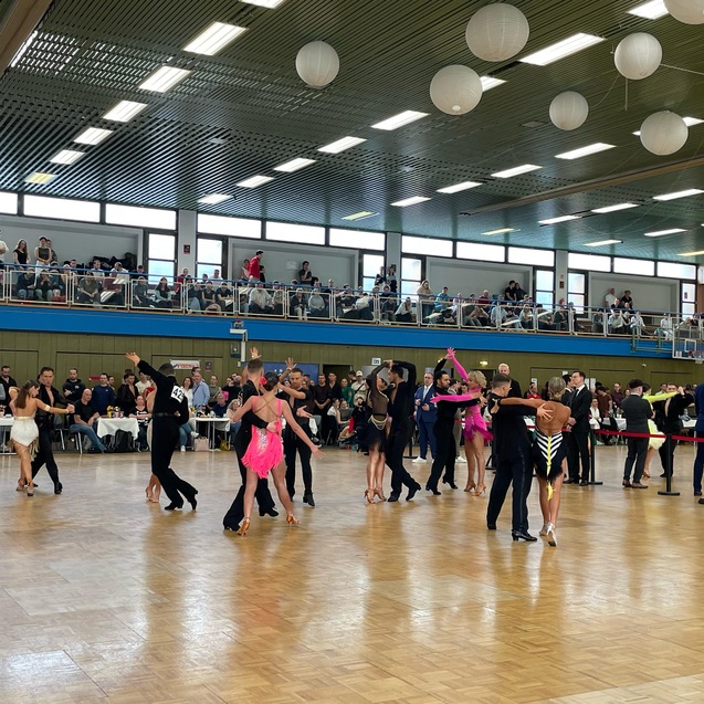 Tanzpaare in verschiedenen Outfits zeigen einen Wettbewerb in einem gro&szlig;en Saal mit Zuschauern auf der Trib&uuml;ne.