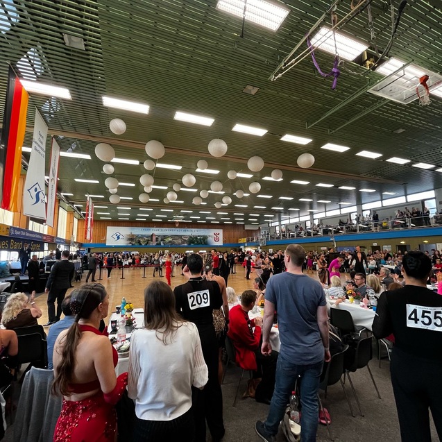 Tanzwettbewerb in einer gro&szlig;en Halle mit Zuschauern, T&auml;nzern und festlich eingedeckten Tischen im Hintergrund.