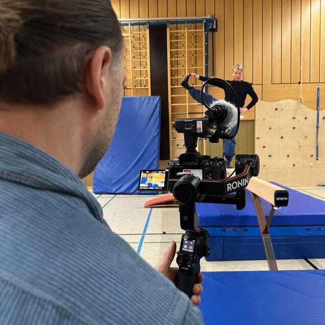 Person filmt eine Gymnastik&uuml;bung in einer Turnhalle mit Kamera auf einem Stabilisator und einem Balancebalken im Vordergrund.