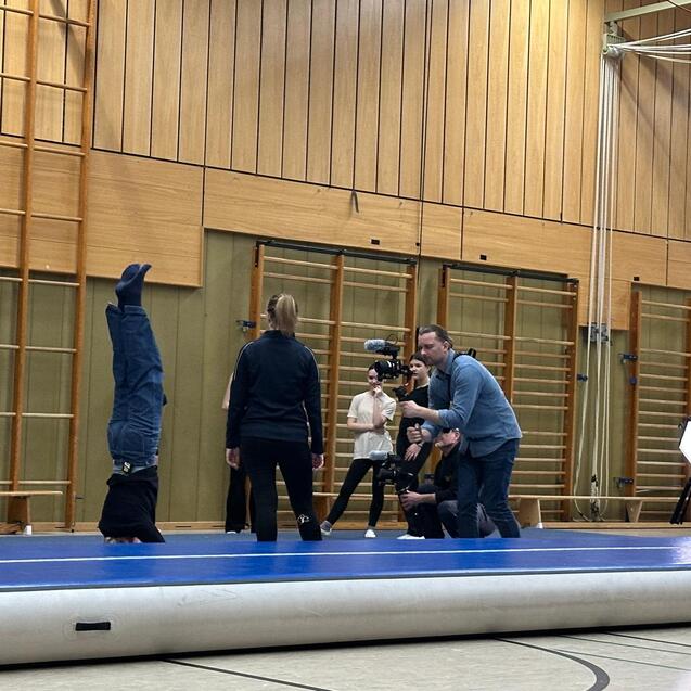 Gruppen&uuml;bung in einer Sporthalle: Zwei Personen machen Handst&auml;nde, w&auml;hrend mehrere Zuschauer das Geschehen beobachten.
