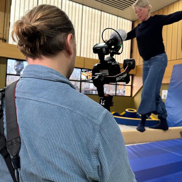 Ein Mann filmt eine Frau, die auf einem Balken in einer Turnhalle balanciert, w&auml;hrend er eine Kamera h&auml;lt.