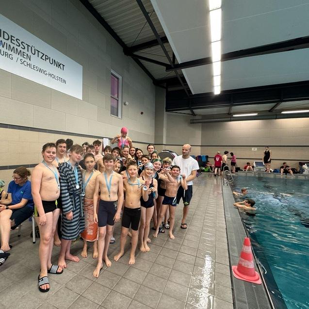 Gruppe von Schwimmern in Badebekleidung mit Medaillen, posieren am Beckenrand eines Hallenbades.