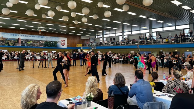 Turnierveranstaltung mit Tänzern in bunter Tanzkleidung, Publikumsbereich und dekorativen Papierlaternen im Hintergrund.