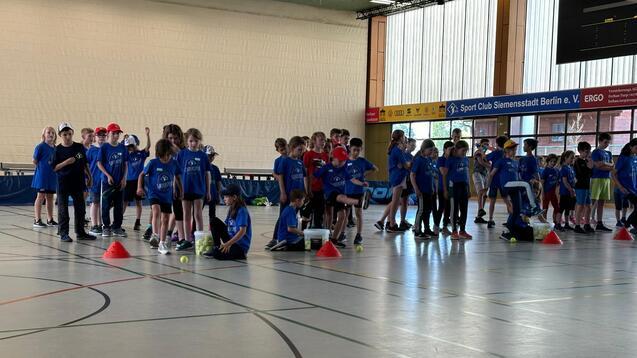 Gruppierung von Kindern in blauen T-Shirts in einer Sporthalle, angereiht an roten Kegeln mit Tennisbällen.
