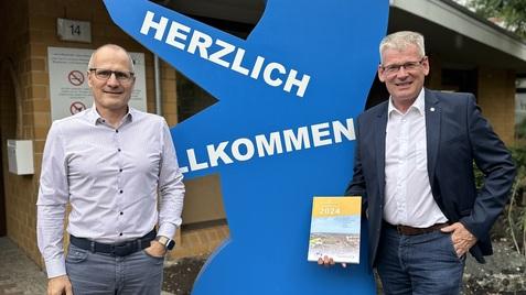 Zwei Männer posieren neben einem blauen Bären mit der Aufschrift „HERZLICH WILLKOMMEN“ vor einem Restaurant.