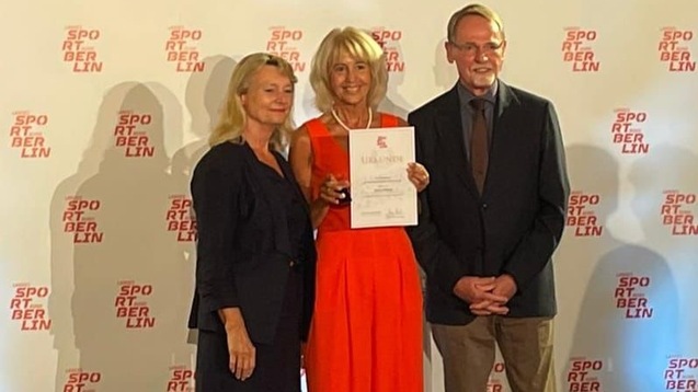 Drei Personen stehen vor einem Hintergrund mit dem Logo "Sport Berlin" und halten eine Urkunde in der Hand.