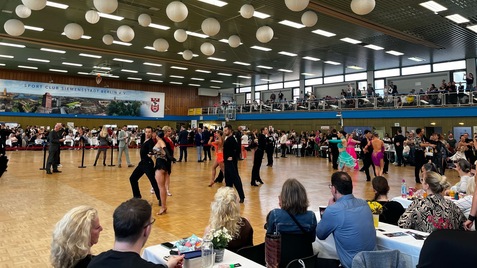 Turnierveranstaltung mit Tänzern in bunter Tanzkleidung, Publikumsbereich und dekorativen Papierlaternen im Hintergrund.