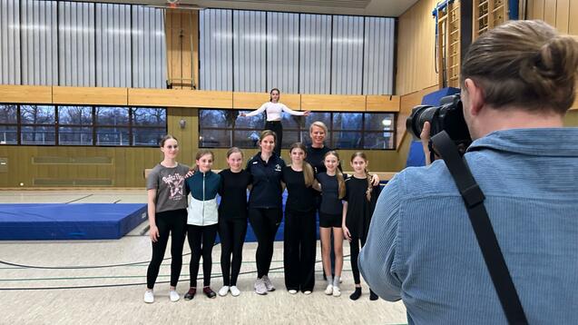 Gruppe von sechs Sportlerinnen in Sportkleidung posiert in einer Turnhalle, während ein Fotograf ein Foto aufnimmt.
