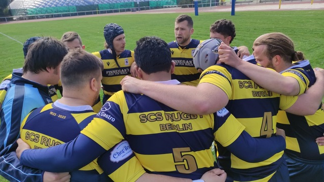 Rugby-Mannschaft in schwarz-gelben Trikots bildet einen Kreis auf dem Spielfeld und bespricht sich vor dem Spiel.