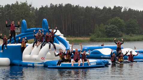 Gruppierung von Personen in Schwimmwesten, die auf schwimmenden Plattformen an einem See spielen und winken.