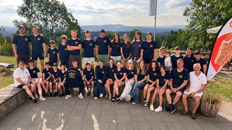 Gruppenfoto von 24 Personen in blauen T-Shirts, im Freien mit Bäumen und Blick auf die Landschaft.