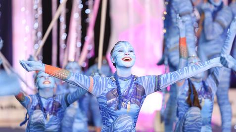 Tanzende Performer in blauen Kostümen mit Gesichtsbemalung, umgeben von festlicher Dekoration und Lichtern auf der Bühne.
