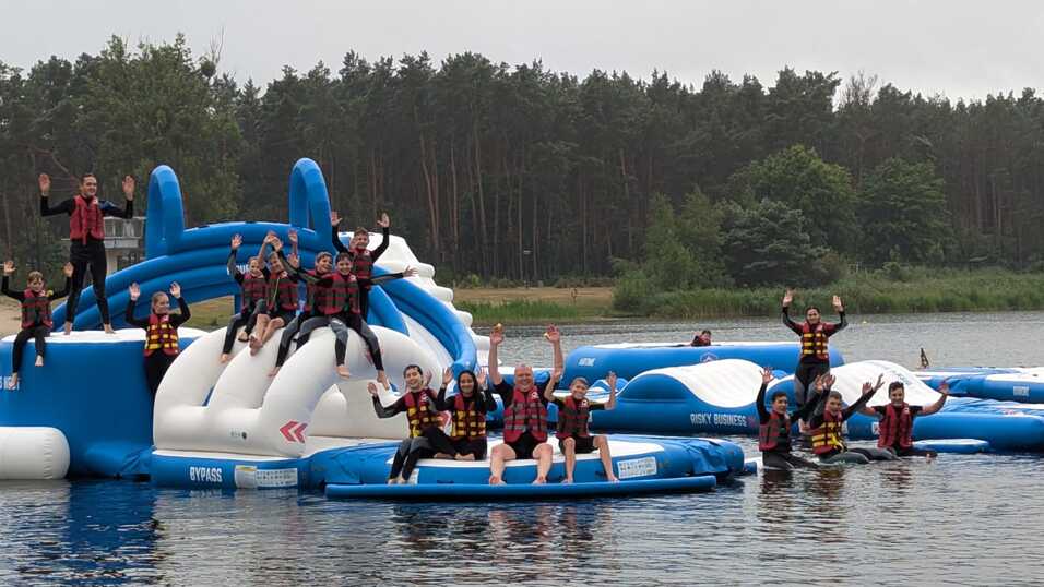 Gruppierung von Personen in Schwimmwesten, die auf schwimmenden Plattformen an einem See spielen und winken.