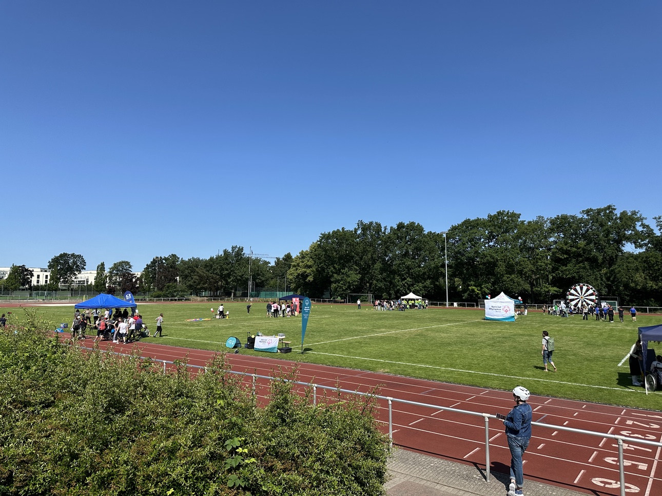 Sportveranstaltung auf einem grünen Feld mit Ständen, Besuchern und einem Glücksrad vor klarem Himmel.