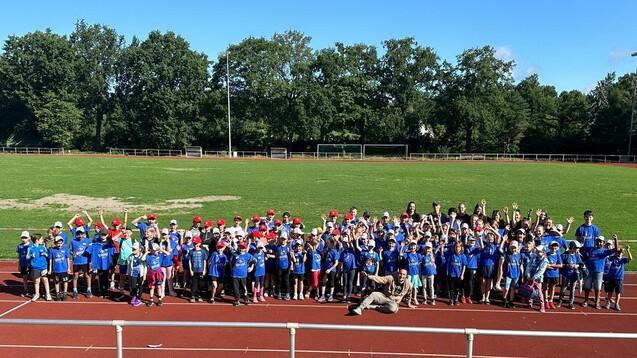 Gruppierung von Kindern in blauen T-Shirts und roten Kappen auf einer Laufbahn, umgeben von B&auml;umen und einem gr&uuml;nen Feld.