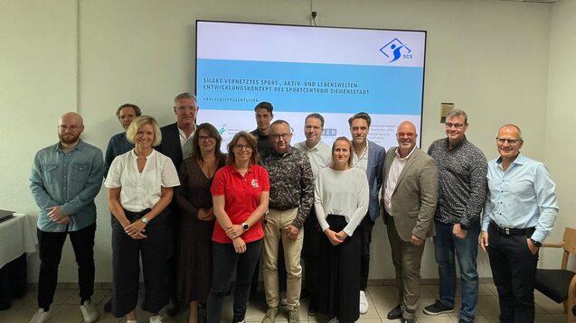 Gruppenfoto von 14 Personen bei einer Präsentation zu einem Sportentwicklungsprojekt in einem modernen Raum.