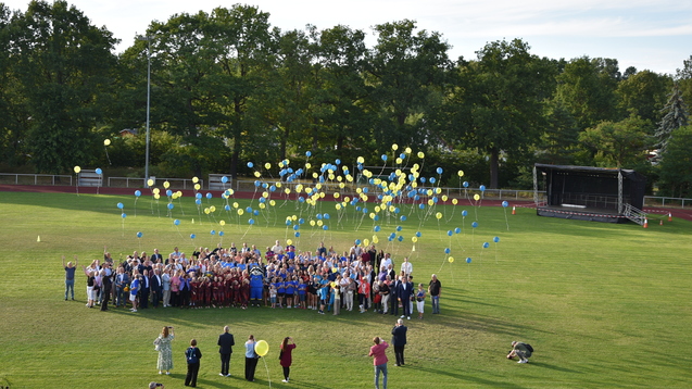 Gruppierung von Menschen auf einem Sportplatz, umgeben von schwebenden gelben und blauen Ballons.