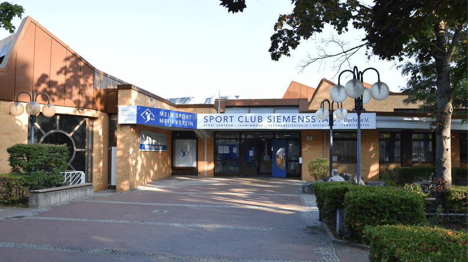 Eingang des Sport Clubs Siemensstadt mit modernen Lampen und Begr&uuml;nung auf dem Gehweg.