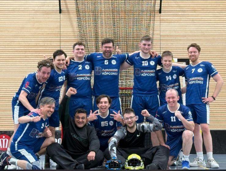 Gruppennfoto einer Handballmannschaft in blauen Trikots, lächelnd und posierend in einer Sporthalle.