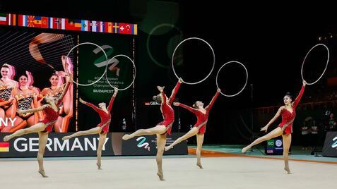 Fünf weibliche Turnerinnen in roten Kostümen synchronisieren Bewegungen mit Reifen auf einer Gymnastikfläche.