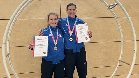 Zwei Athletinnen in blauen Sportjacken stehen in einem großen Holzring und halten Medaillen und Urkunden in den Händen.
