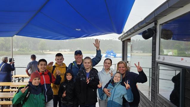 Gruppe von acht Personen steht unter einem blauen Pavillon an einem Gewässer, einige zeigen Victory-Zeichen.