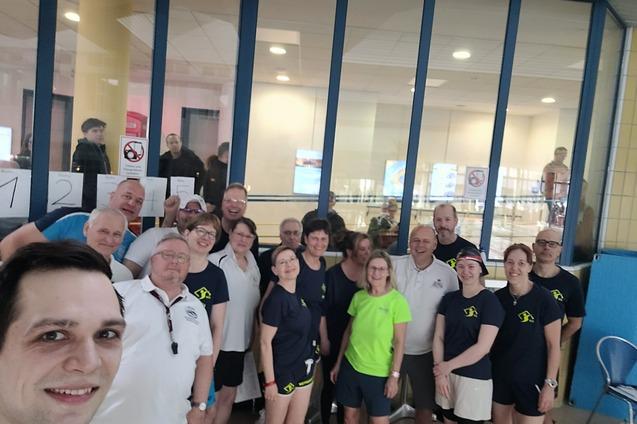 Gruppe von 18 Personen in Sportkleidung posiert vor einer Fensterfront in einem Schwimmbad.