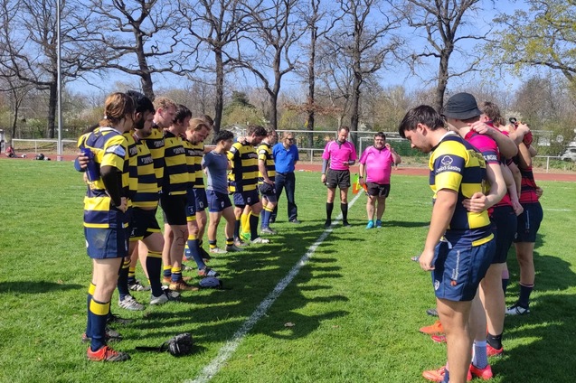 Zwei Rugby-Teams in gestreiften Trikots stehen auf einem Feld, w&auml;hrend Schiedsrichter und Zuschauer beobachten.