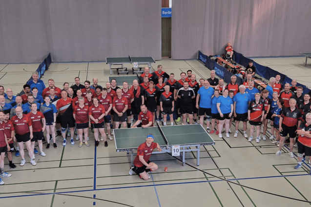 Gruppenfoto von Tischtennisspielern in Sportkleidung auf einer Sporthalle mit Tischtennisplatten im Hintergrund.
