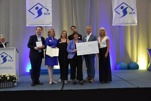 Gruppenszene mit sieben Personen bei einer Preisverleihung, einige halten einen Scheck und eine Urkunde.