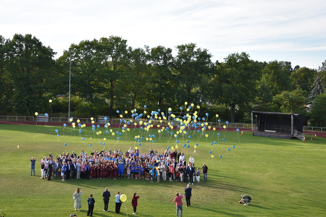 Gruppierung von Menschen auf einem Sportplatz, umgeben von schwebenden gelben und blauen Ballons.